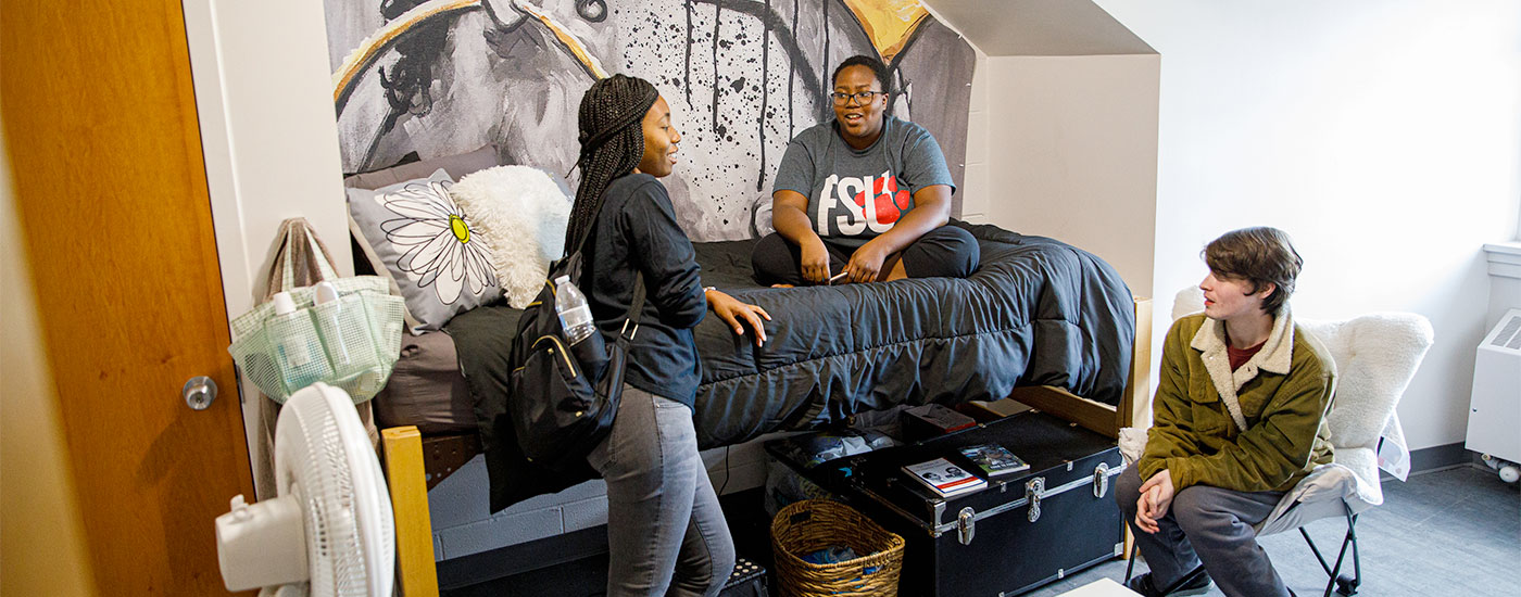 Students chat in their dorm room. One student is sitting on the bed, one is standing, and one is sitting in a chair.